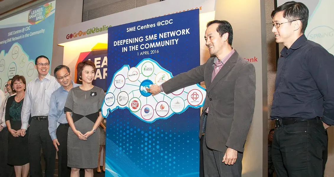 A group of people stands near a large board with the text "Deepening SME Network in the Community" and connected logos.